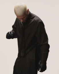 Blonde male model wearing a dark collared shirt and black leather gloves looking down against a neutral studio background.
