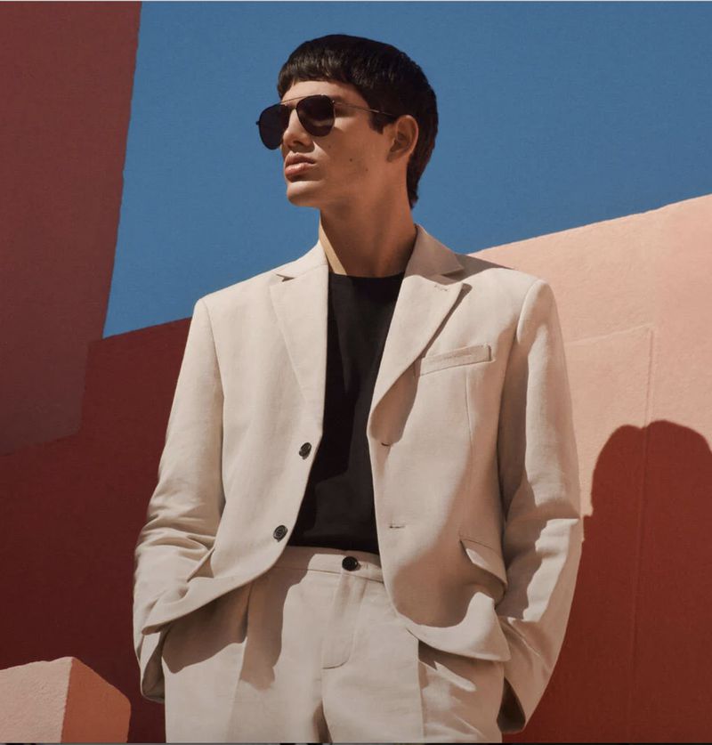 Man poses in a light suit and sunglasses against a wall with blue sky. Fashion shot shows stylish menswear in a vibrant outdoor setting.