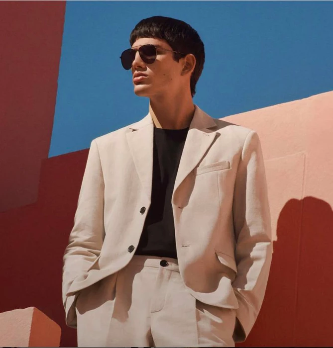 Man wearing a cream blazer and sunglasses posing against pink architectural walls and a blue sky.