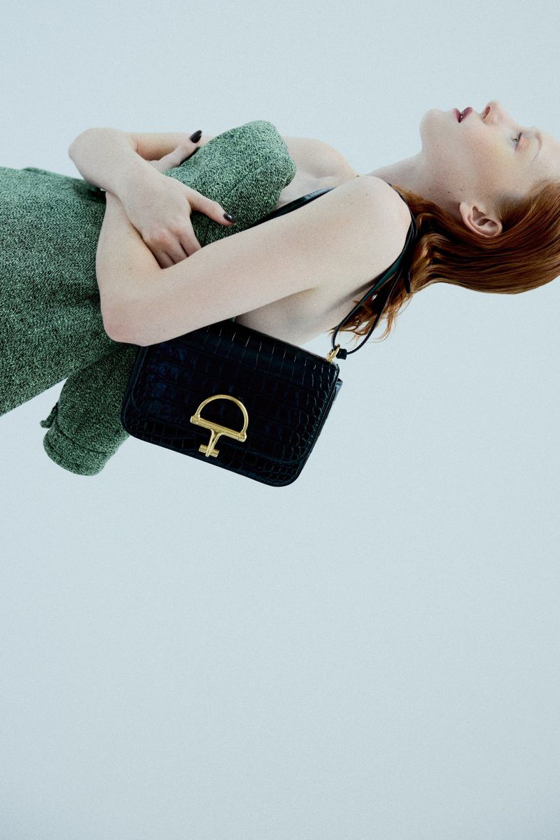 Woman with red hair lying down in a light green sweater with a black leather purse with a gold closure on a white background.