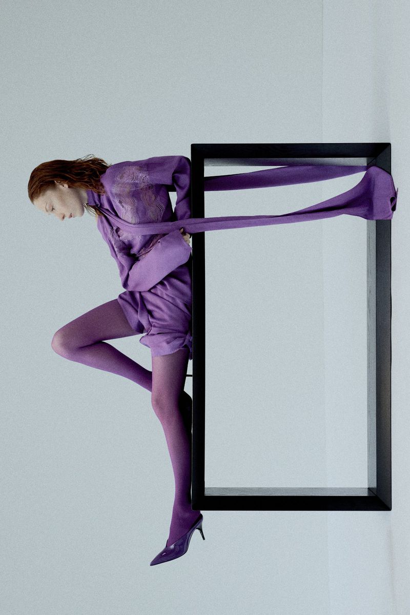 Fashion editorial image featuring a red-haired woman in a lilac outfit, posing in a black picture frame against a clean white background.