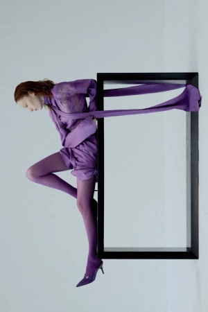 Fashion editorial image featuring a red-haired woman in a lilac outfit, posing in a black picture frame against a clean white background.