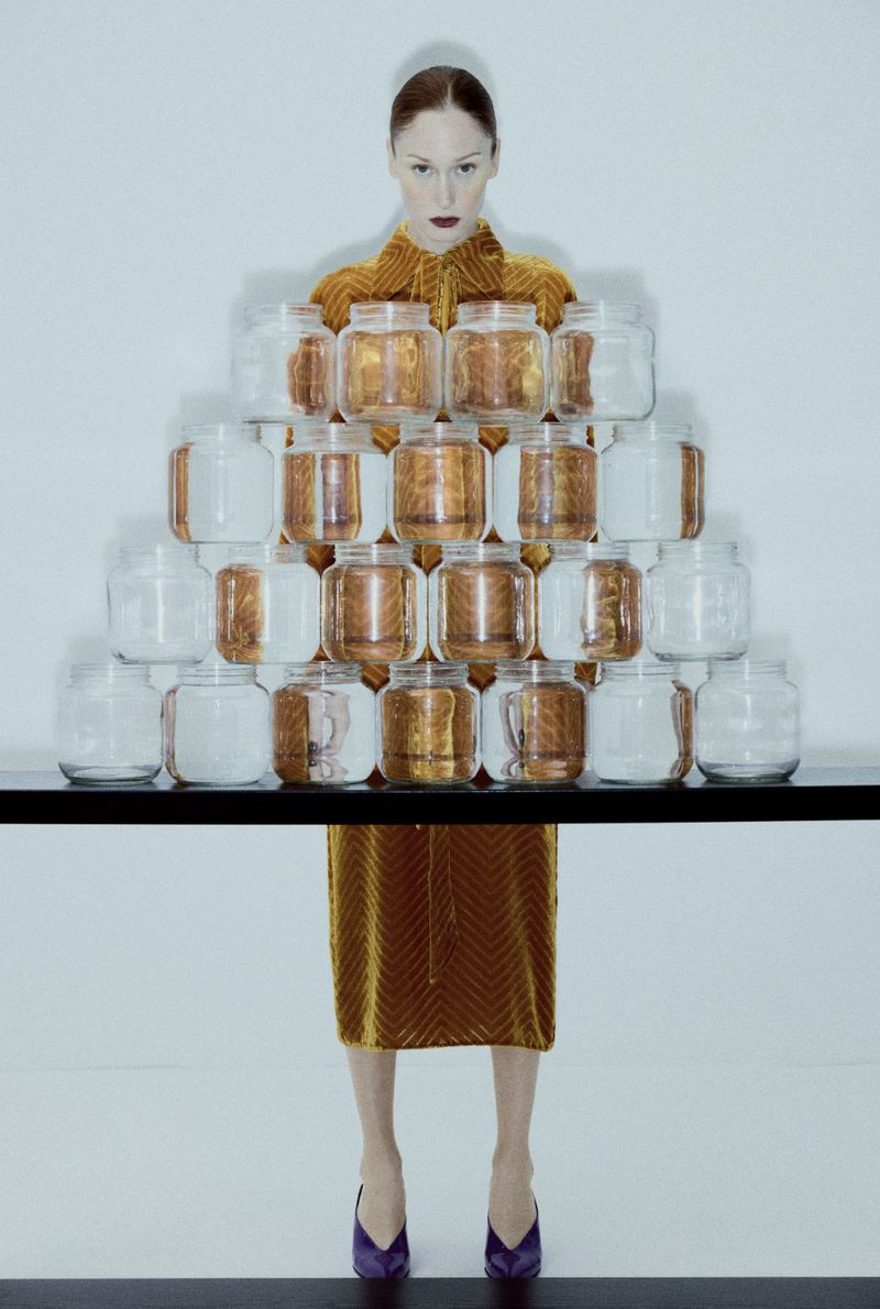Fashion photography featuring a slender woman in an amber velvet dress and purple shoes, posing with a pyramid of glass jars.