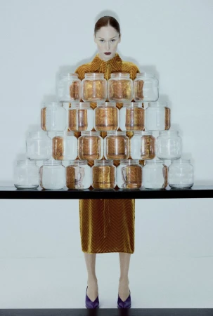 Fashion photography featuring a slender woman in an amber velvet dress and purple shoes, posing with a pyramid of glass jars.