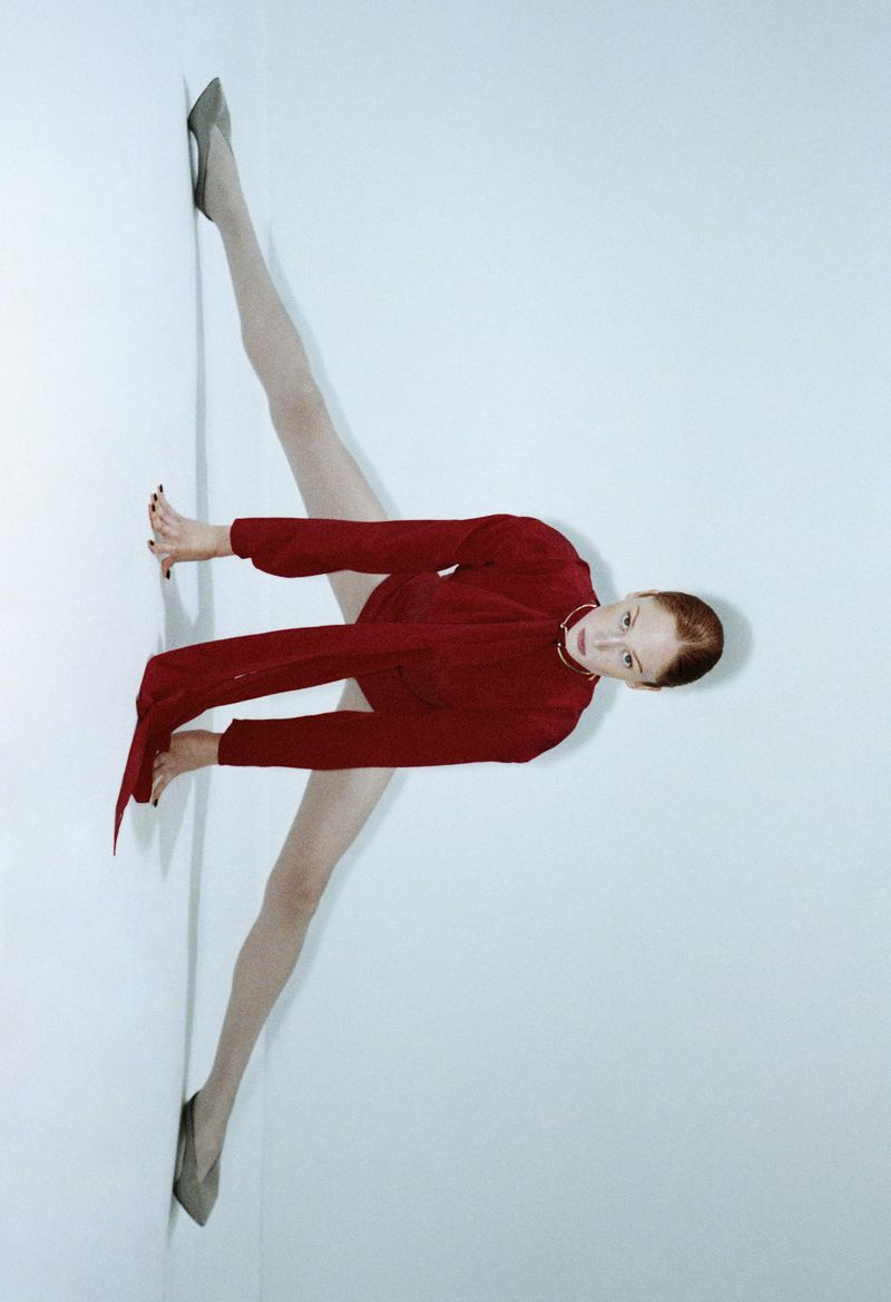 Adult woman in red long-sleeve top and gray shoes performing a split on a white wall, fashion photography, full shot.