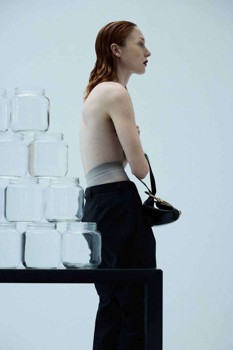 Model strikes pose on black table next to pyramid of glass jars in studio; black trousers, bare torso, wet red hair and black handbag.