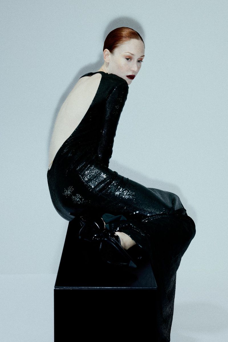 Woman in a long black sequin dress poses while seated on a black cube in a studio setting with a light gray gradient backdrop.