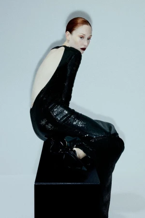 Woman in a long black sequin dress poses while seated on a black cube in a studio setting with a light gray gradient backdrop.