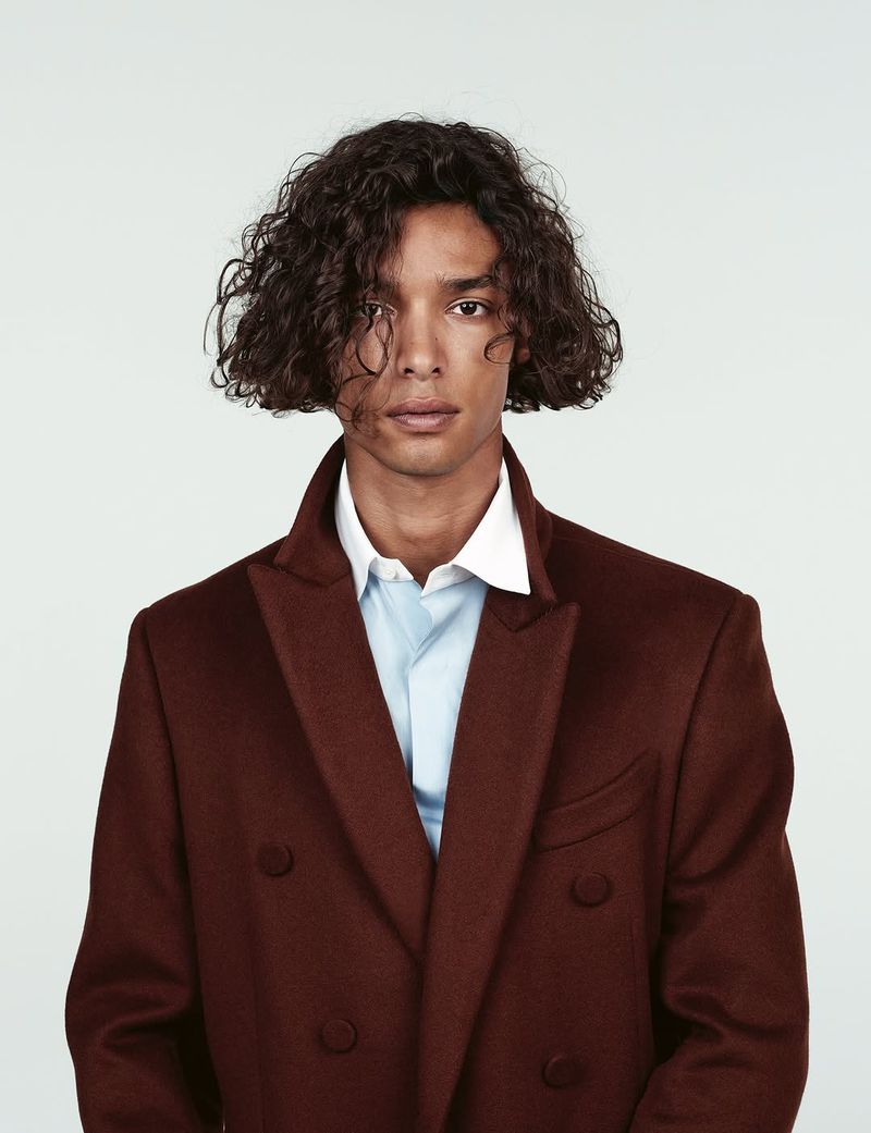 Adult male model with dark curly hair, wearing a burgundy overcoat and a light blue collared shirt in a studio fashion shot.