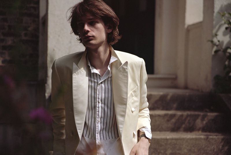 Portrait of a young adult male wearing a cream-colored suit, standing on the steps of a building in an outdoor setting.