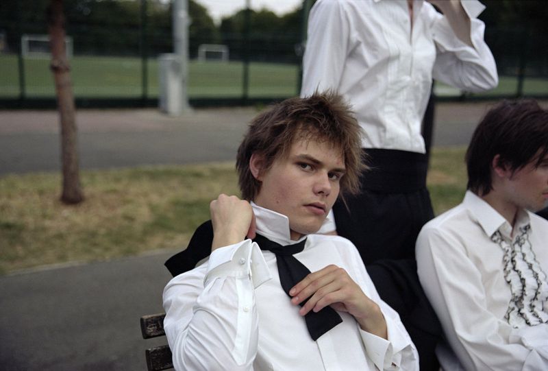 Three young adults in white shirts and black bow ties sit on a bench in an outdoor park setting, captured in a medium shot.