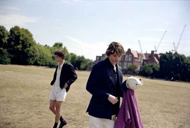 Two young adult men in jackets and casual pants stand on a grassy field under a bright sky, with buildings and cranes in the distance.