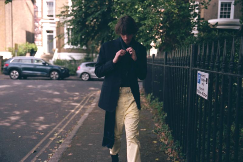A stylish adult male walks on a city sidewalk, wearing a black jacket, yellow pants, and black boots, with cars and buildings visible.