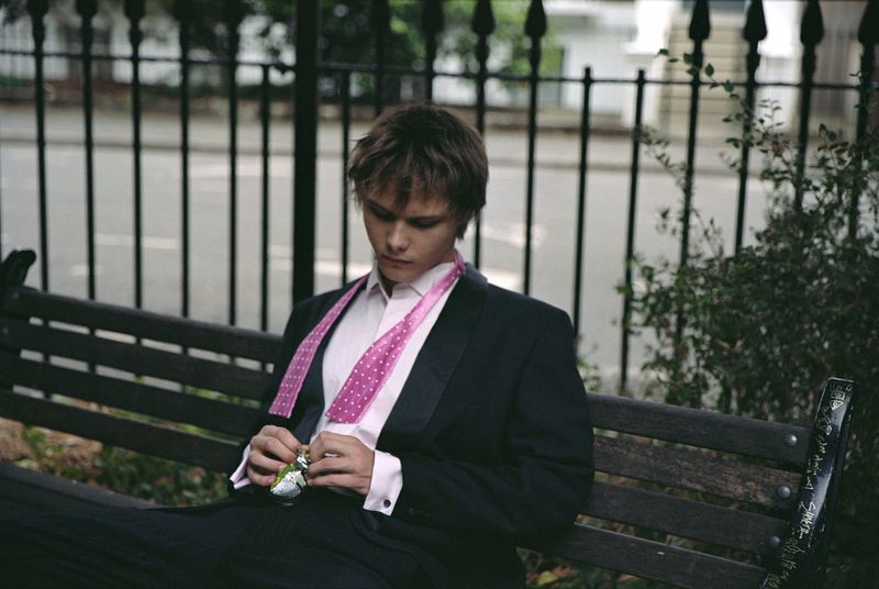 Young adult male in a black suit and pink polka dot tie sits on a bench outdoors, looking down and handling a small object in his hands.
