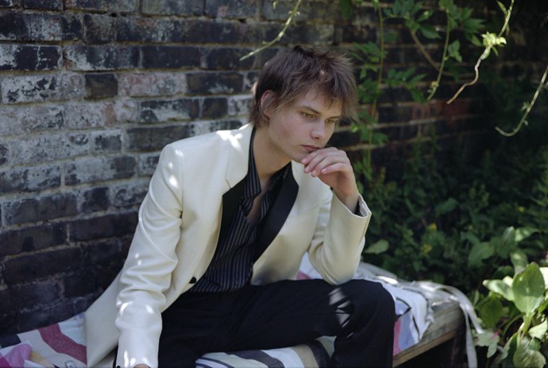 Young adult in off-white suit sitting outside on striped cushion, hand to chin, looking away from camera.