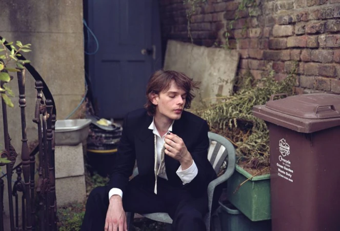 A young adult male in a black suit sits in an outdoor setting with brick walls and greenery, holding a cigarette and wearing an unbuttoned shirt.