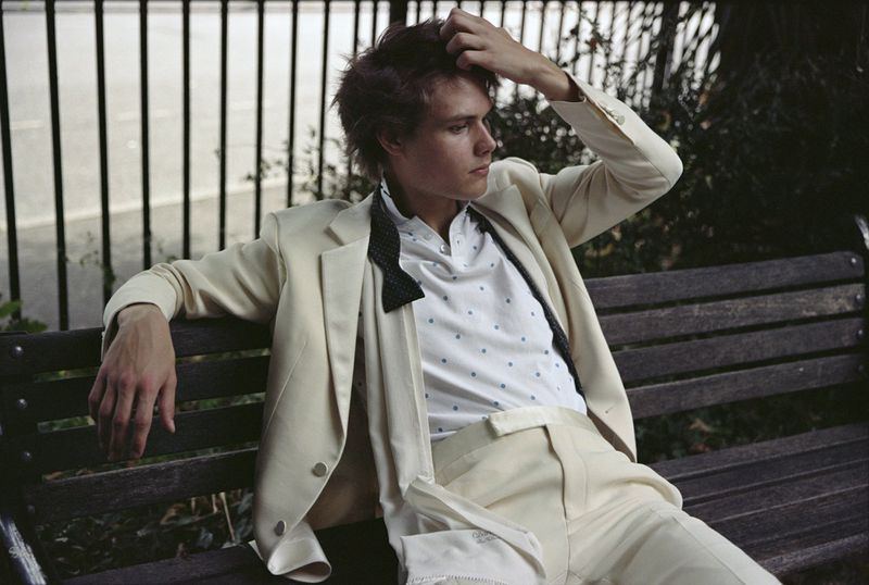 Young adult male in a white suit sitting on a park bench with a black bow tie and polka dot shirt with trees and a fence in the background.