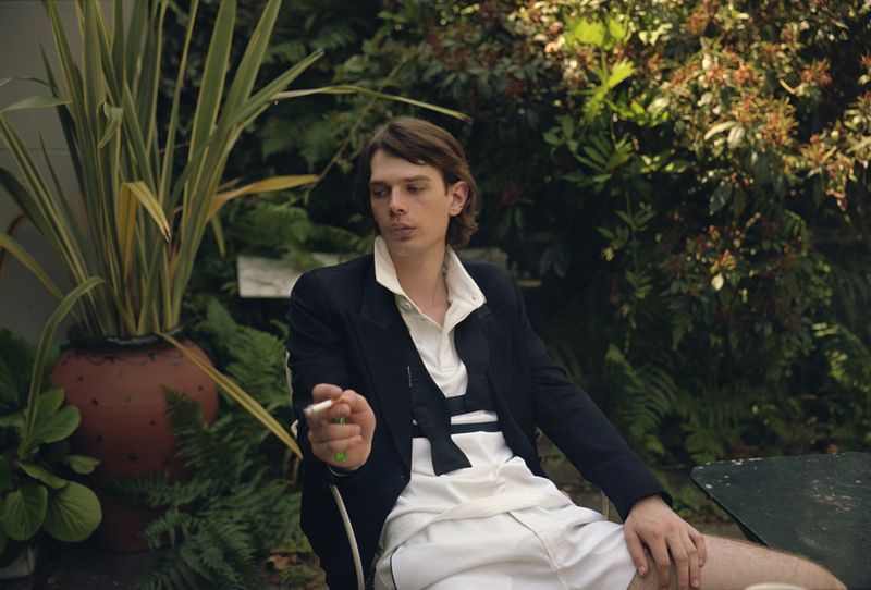 Man in blazer and shorts sits outside in lush greenery, smoking a cigarette; natural light, relaxed pose, medium shot, plants in background.