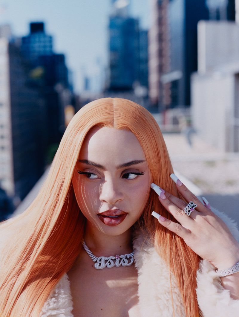 Portrait of an adult woman with long orange hair, wearing statement jewelry and a white jacket against an urban city backdrop.