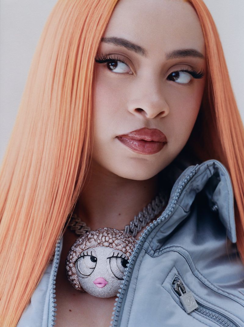 Headshot of a female with long orange hair and a silver jacket. She is wearing a doll head pendant. Shot in a photo studio with white backdrop.