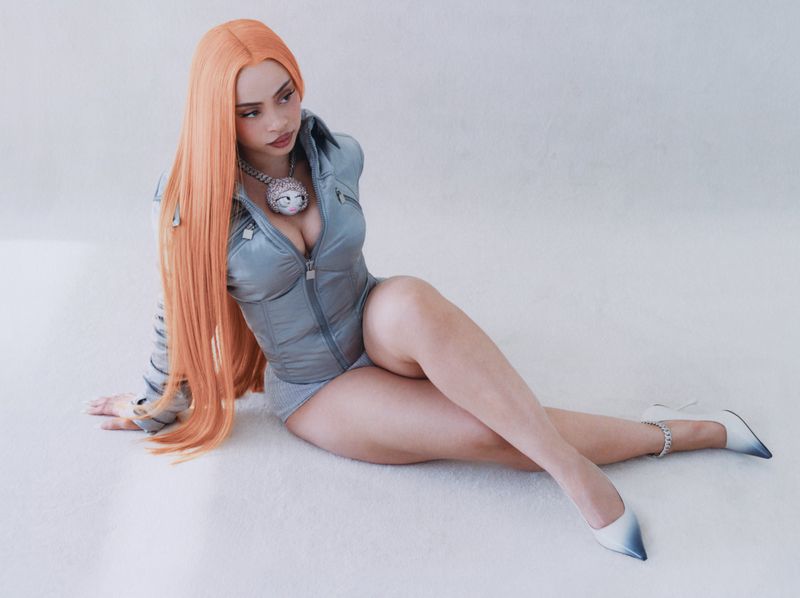 Woman with orange hair sits on a white surface wearing a gray, puffy zip-up top and heels in a studio setting, looking to the side.