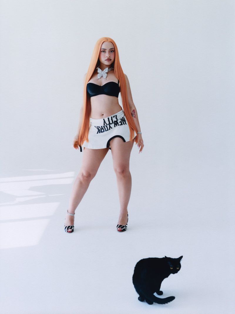 A model with long orange hair, wearing a black top and white skirt, stands in a white studio with a black cat at her feet.