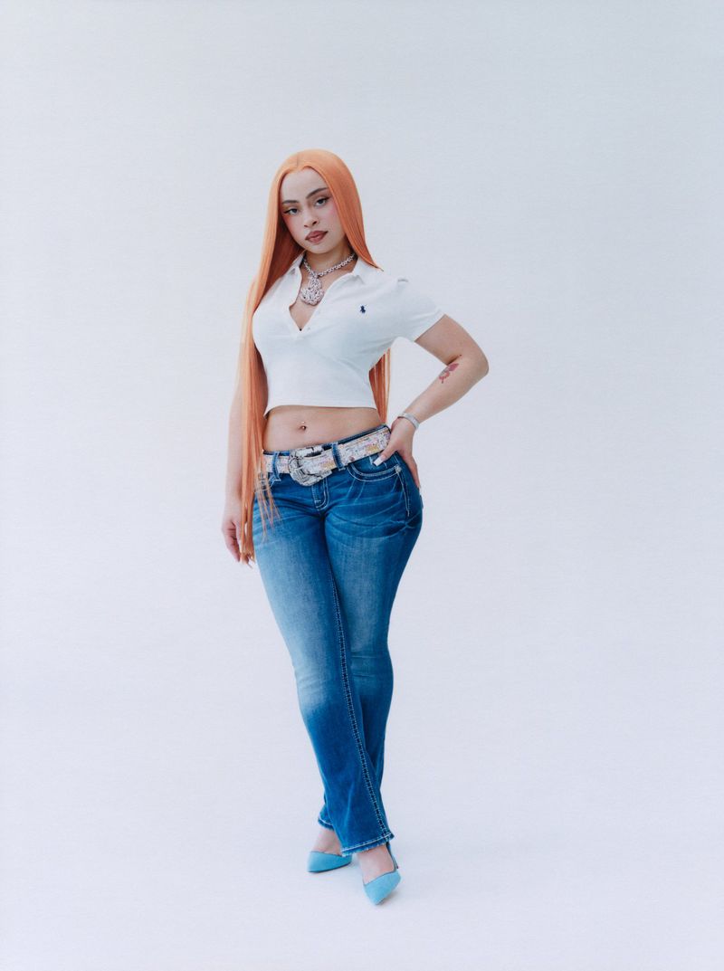 Full-length shot of a woman with long orange hair, wearing a white cropped shirt and blue jeans, posing against a white backdrop.