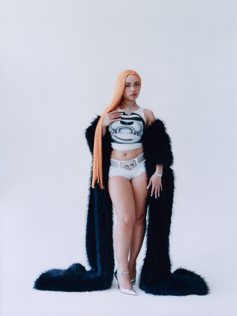 A woman with red hair is posing in a photo studio, wearing a white top, white shorts, and white heels, accessorized with jewelry.