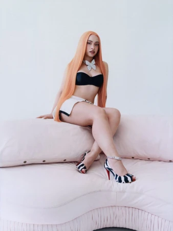 A fair-skinned adult female with long orange hair, wearing black lingerie and a white skirt, sits on a pink couch in a studio.