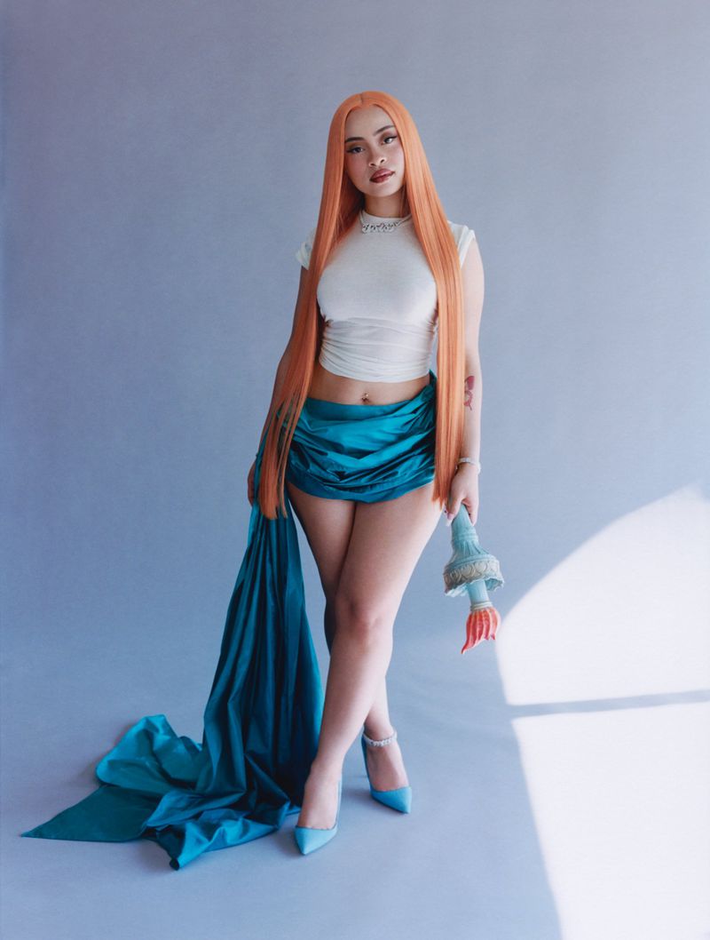 Full-length portrait of a young woman with orange hair wearing a white top and blue skirt, holding a toy against a gradient blue background.