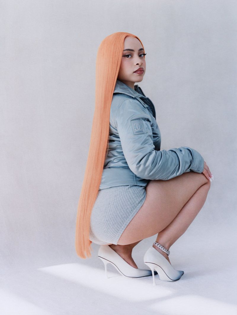 Portrait of a woman with long peach-colored hair squatting in a light blue jacket, matching shorts, and white high heels in a studio.