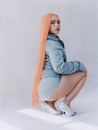 Portrait of a woman with long peach-colored hair squatting in a light blue jacket, matching shorts, and white high heels in a studio.