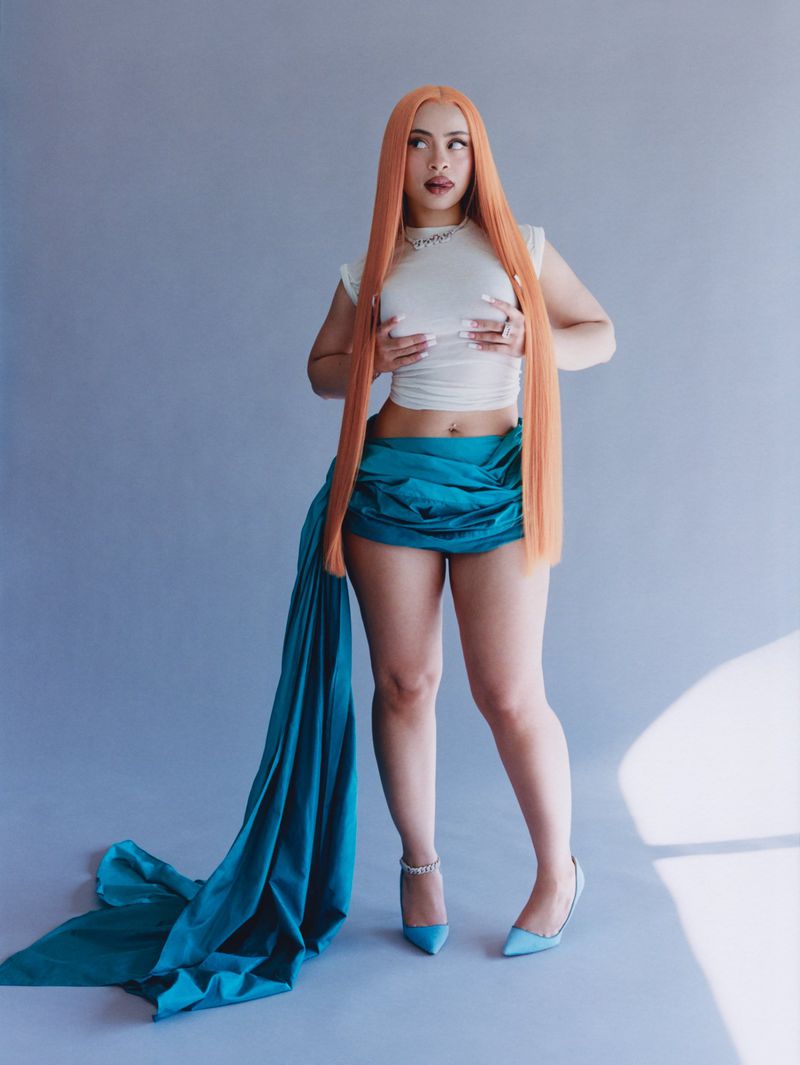 Full-body shot of a woman with long red hair, wearing a white top and teal skirt, posing in front of a light blue studio backdrop.
