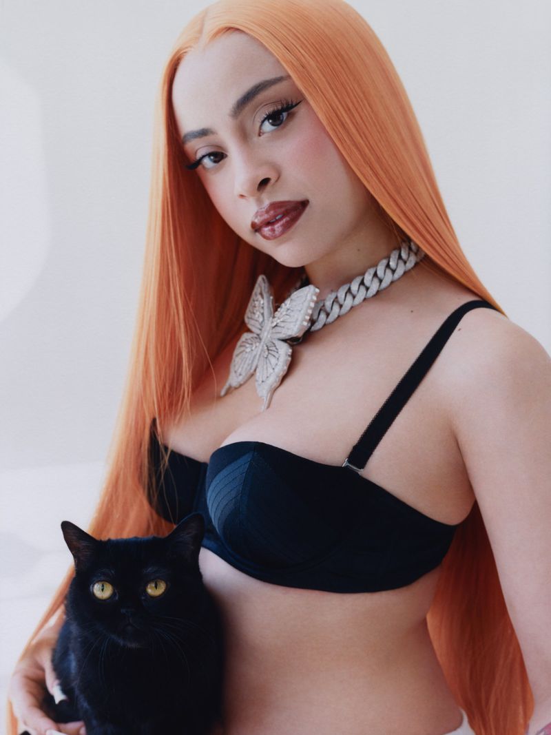 A woman with long orange hair poses with a black cat. She is wearing a black bra and silver necklace with a butterfly pendant.