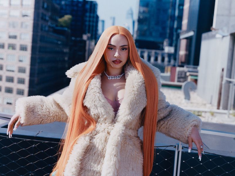 Fashionable woman with orange hair and fur coat poses against a cityscape backdrop, capturing a blend of urban style and glamour.