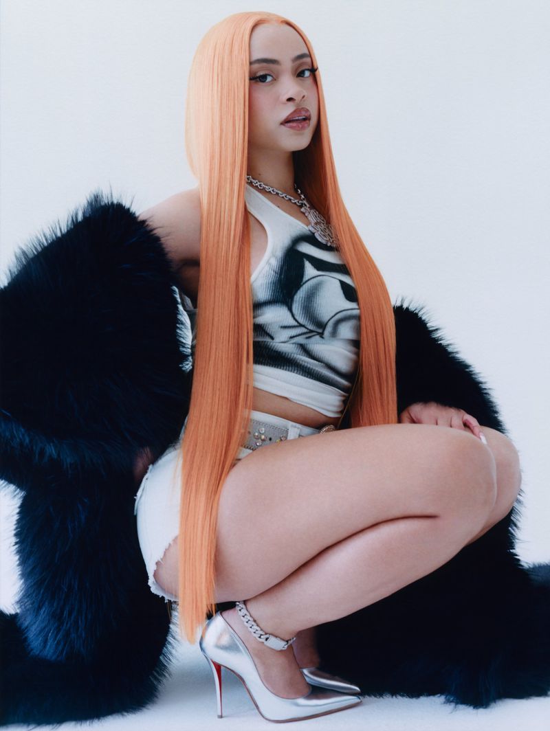 A crouched woman with long orange hair, wearing a white top, shorts, and silver high heels, poses against a white backdrop.