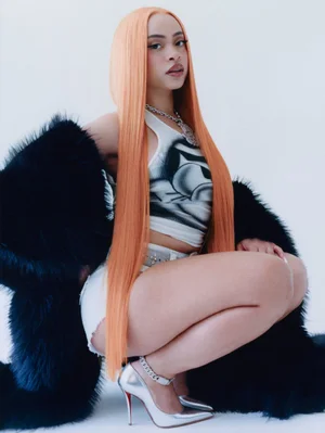 Ice Spice crouching in a white tank top, shorts, silver heels, and oversized black fur coat against a white background.