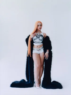 Ice Spice wearing a white graphic crop top and black faux fur coat against a seamless white background