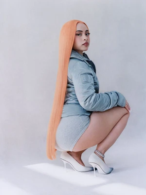 Ice Spice crouching in profile wearing a blue jacket and white heels against a plain light grey studio backdrop