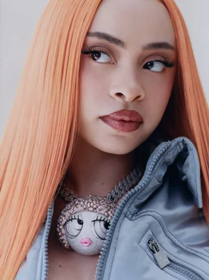 Close-up portrait of Ice Spice with long orange hair wearing a grey jacket and large cartoon pendant necklace against a plain background.