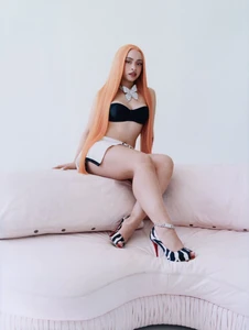 Ice Spice with long orange hair wearing a black strapless top seated on a pink sofa