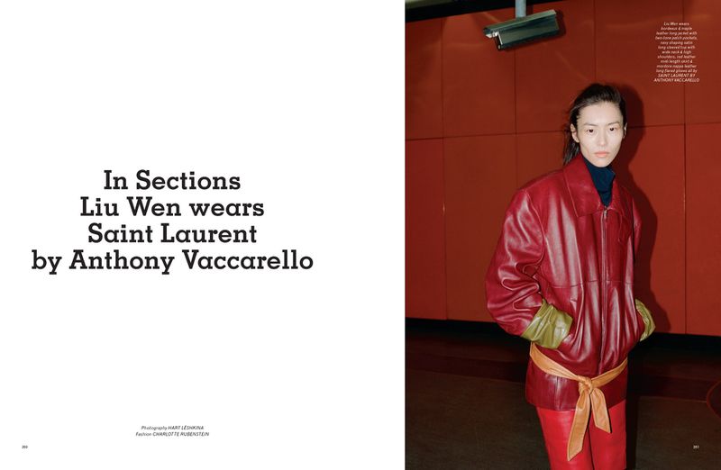 Editorial fashion portrait of a woman wearing a red leather jacket and pants, standing indoors against a red wall with a security camera.
