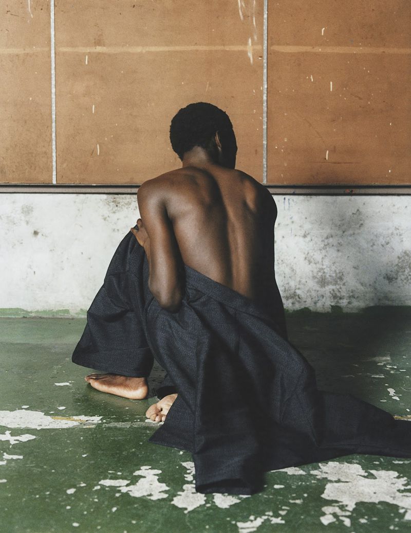 Back view of an adult with short hair sitting on a distressed green floor, wearing dark flowing pants in front of a brown paneled wall.