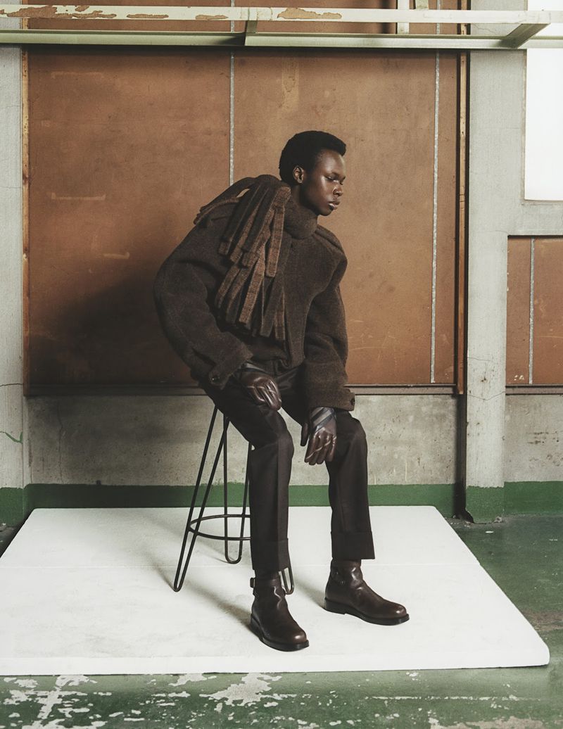 Young black man models in a brown jacket, matching pants, and boots, sitting on a black stool in a minimalist studio with a neutral palette.
