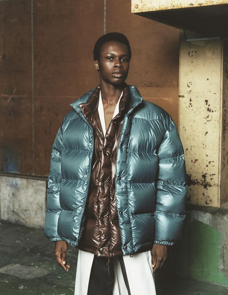 Man in puffy teal down jacket and white pants poses in front of an industrial wall. He is looking straight at the camera.