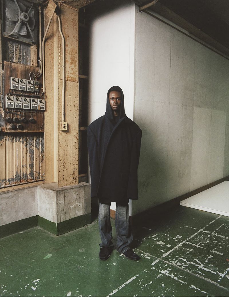 Fashion shot of a Black adult male in a dark oversized coat, blue jeans, and black shoes, standing in a grungy industrial-style indoor setting.