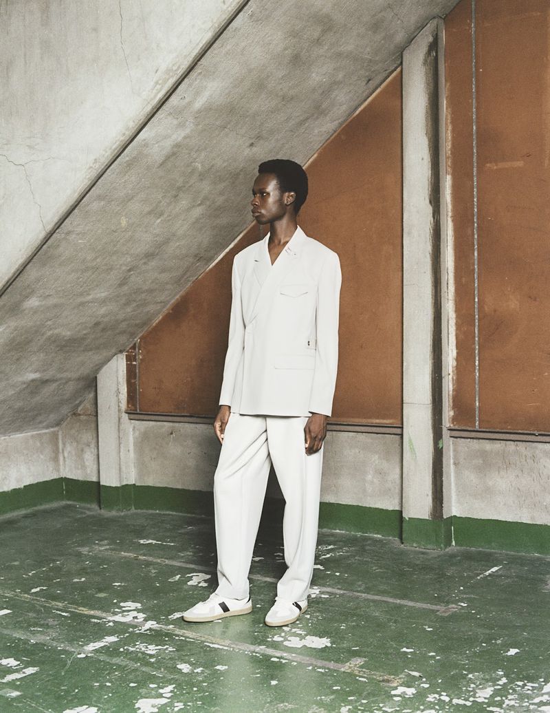 Fashionable Black man in a suit stands indoors with a concrete ceiling and a green floor, exuding cool style.