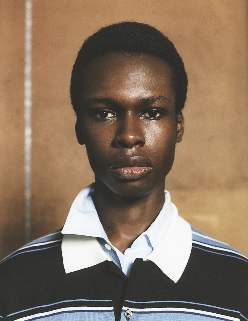 Portrait of a young Black man wearing a striped polo shirt. The photo features natural skin tones and earth-toned background, focusing on his face.