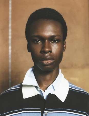 Portrait of a young Black man wearing a striped polo shirt. The photo features natural skin tones and earth-toned background, focusing on his face.