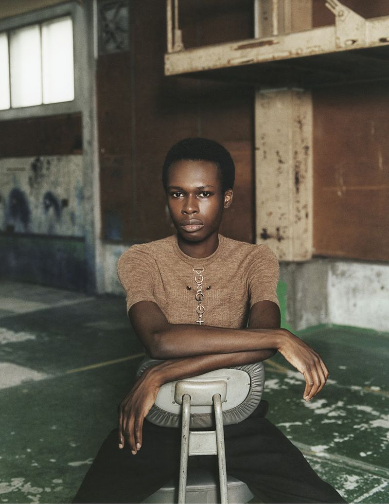 Indoor portrait of an African-American person with short hair, wearing a brown top, sitting on a grey chair in a weathered industrial space.